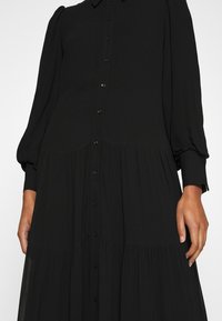 Robe noire à manches longues boutonnée avec jupe à volants et manches bouffantes, portée par une personne les mains détendues sur les côtés.