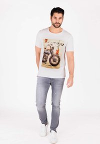 Maglietta bianca a maniche corte con una grafica di motocicletta stampata sul fronte, abbinata a jeans grigi chiari strappati e sneakers bianche.