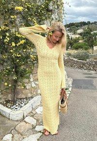 Γυναίκα με μακρύ κίτρινο πλεκτό φόρεμα στέκεται κοντά σε ανθισμένο κλήμα σε πέτρινο τοίχο, κρατά μια πλεκτή τσάντα και αγγίζει ένα κίτρινο λουλούδι στα μαλλιά της.