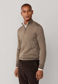 Bruine gebreide sweater met kwart-rits, gemaakt van zacht materiaal, met een geribde zoom en manchetten, gedragen over een witte overhemd met kraag.