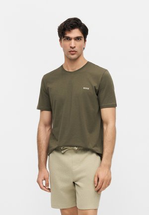 Man met donker haar die een olijfgroene BOSS T-shirt en beige short met trekkoord draagt, staat voor een effen lichte achtergrond.