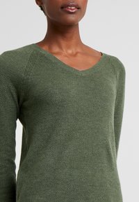 Maglione verde lavorato a maglia con scollo a V, caratterizzato da un tessuto testurizzato e maniche raglan senza cuciture, che mette in risalto un design morbido e traspirante.