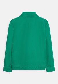 Grøn langærmet jakke med opretstående krave, synlig syning ved manchetter og kant, vist fra bagsiden på en hvid baggrund.