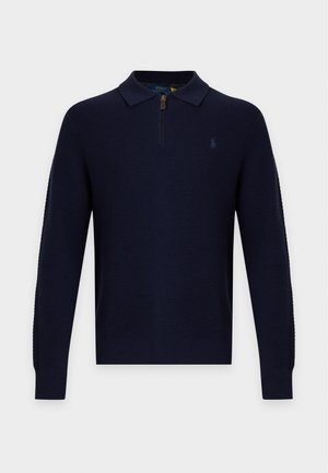 Homme portant un pull bleu marine foncé avec col zippé, pantalon assorti et baskets blanches, debout avec une main dans la poche.