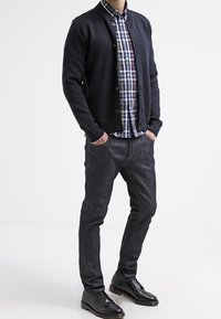 Marineblaue Strickjacke mit Knöpfen, kariertes Hemd in Blau, Grün und Weiß, dunkle Jeans aus Denim und schwarze Lederschuhe, stehend vor einem neutralen Hintergrund.