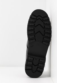 Semelle de botte noire avec des crampons profonds pour la traction, présentée verticalement sur un fond d'angle blanc.