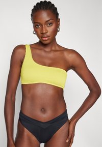 En-skulder gul bikini topp med teksturert stoff og bølgete kanter, kombinert med høy-sydd svart bikini bukser med overlappende design.