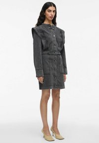 Femme portant une robe en jean noire à boutons avec des épaules structurées et des chaussures à talons beiges, se tenant devant un fond blanc.