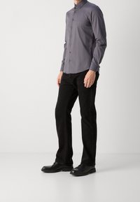 Camisa de mangas largas abotonada en gris oscuro con un corte ajustado, combinada con jeans negros y zapatos negros. La camisa tiene un logotipo sutil.