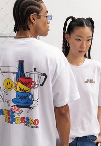 White cotton t-shirts featuring colorful graphic prints: espresso cups, a coffee pot, a smiley face, and the text “MORE ESPRESSO”.