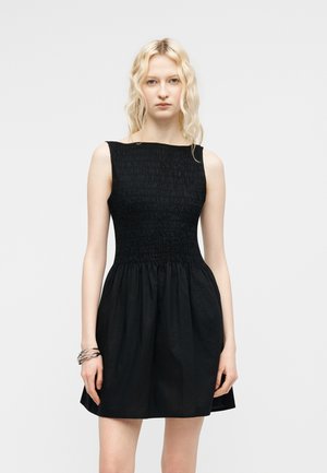 Ung kvinde med bølget blondt hår iført en ærmeløs sort kjole med smock-øjeblik og udvidet nederdel, stående mod en ensfarvet baggrund.