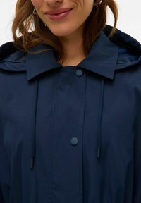 Mörkblå regnjacka med stor krage, tryckknappar, justerbar dragsko och slät yta. Fokus på funktionalitet och minimal design.