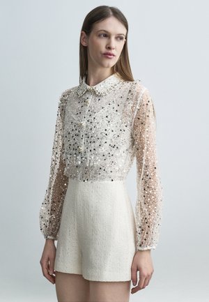 Donna che indossa una maglia trasparente a maniche lunghe con paillettes argentate e pantaloncini bianchi strutturati a vita alta, in piedi davanti a uno sfondo grigio chiaro.