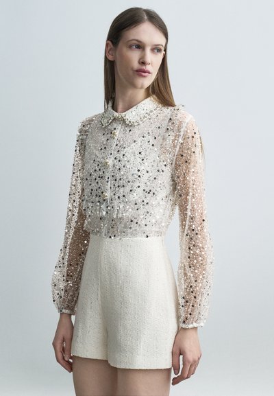 Femme portant une chemise transparente à manches longues ornée de sequins argentés et un short blanc texturé taille haute, debout devant un fond gris clair.