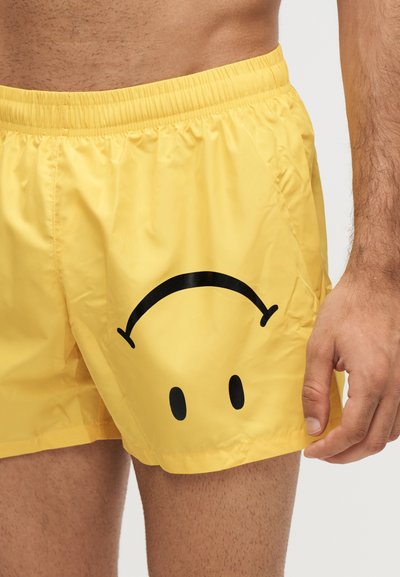 Homme portant un short de bain jaune avec un graphique de visage souriant noir à l’envers sur le côté avant gauche.