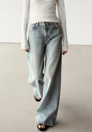 Person trägt hellblaue Jeans mit weitem Bein, ein weißes, eng anliegendes Langarmshirt und schwarze Sandalen mit offenen Zehen und geht auf einem glatten Innenboden.