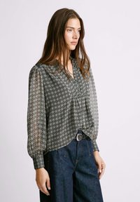 Blusa in bianco e nero a fantasia con maniche lunghe a palloncino, un piccolo colletto e parte anteriore arricciata. Indossata con pantaloni in denim scuro a vita alta.