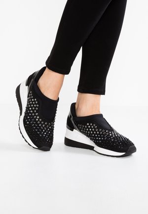 Stopy noszące czarne, wsuwane sneakersy na koturnie z srebrnymi ćwiekami, zestawione z czarnymi dopasowanymi spodniami na białym tle.