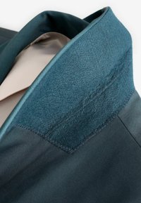 Collar de blazer verde oscuro con una tela de fieltro texturizada, con un acabado de satén suave por debajo. Detalle de costura a lo largo del borde.