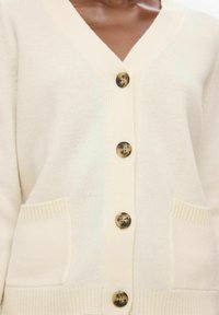 Cárdigan de punto crema con escote en V, bolsillos frontales y cinco grandes botones marrones con un diseño veteado. Textura suave. Tejido duradero.
