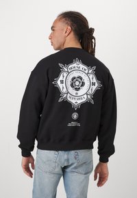 Hanorac negru cu un grafic alb mare pe spate, care prezintă detalii florale și text intricate, cu manșete și tiv texturate. Jeans albastru deschis.