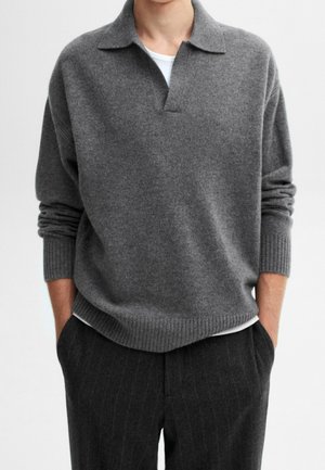 Uomo con indosso un maglione grigio lavorato a maglia con colletto e una maglietta bianca sotto, abbinato a pantaloni scuri a righe sottili, con le mani in tasca.