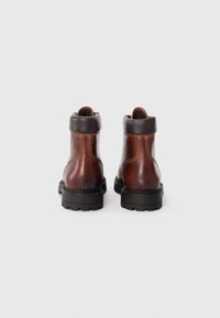 Botas de cuero marrón hasta el tobillo con puntera redondeada, suela de goma negra y puño superior oscuro en contraste. Detalles de costura visibles a lo largo de las costuras.
