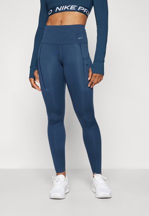 Høj taljede navy blå leggings lavet af strækstof, med sidelommer og en glat tekstur, parret med hvide træningssko.