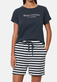Kvinna som bär en marinblå Marc O’Polo t-shirt och marinblå och vita horisontella randiga shorts med dragsko, med händerna i fickorna.