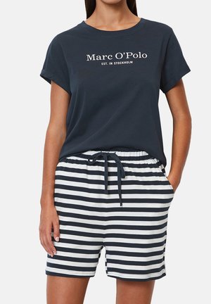 Kvinna som bär en marinblå Marc O’Polo t-shirt och marinblå och vita horisontella randiga shorts med dragsko, med händerna i fickorna.