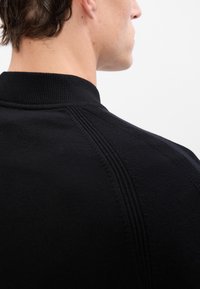 Sort strikket genser med høy krage, strukturert materiale og subtile ribbede detaljer langs skuldrene og halsen.
