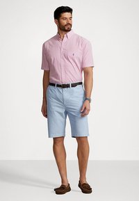 Vīriešu apģērbs, kas ietver sarkanu un baltu svītrotu īsām piedurknēm pogājamu kreklu, gaiši zilos šortus un brūnus laivu apavus. Ievērojama jostas detaļa.