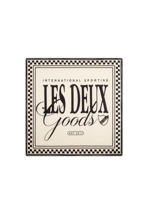Neliölautanen, jossa on kerma tausta ja musta teksti. Ruudullinen reuna kehystää "LES DEUX Goods" ja "EST 2011" pienen logon kera.