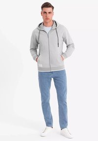 Haut de sweat à capuche zippé gris clair avec cordons, poches avant et ourlet à bande, associé à un jean bleu clair et des baskets blanches.
