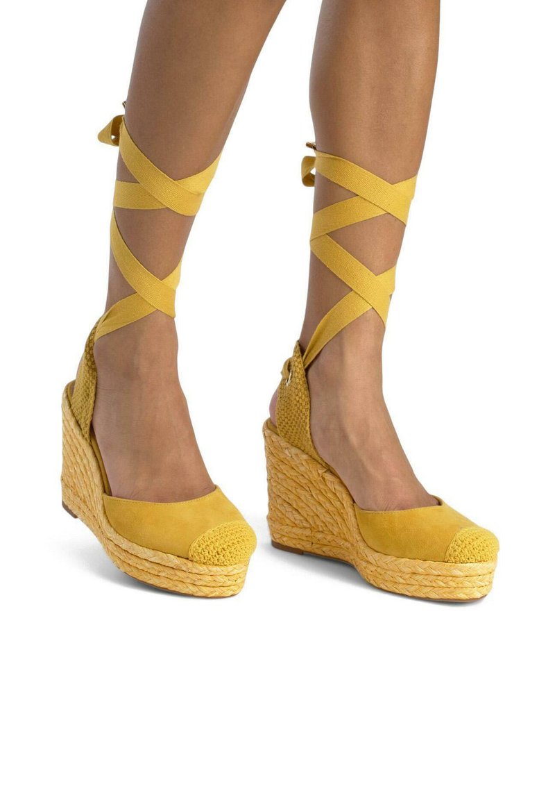 Scarpe con tacco a zeppa gialle in espadrillas con base in paglia testurizzata, tomaia in suede e cinturini incrociati alla caviglia. Design con punta arrotondata e dettaglio di cappuccio intrecciato.