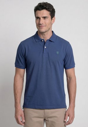 Hombre con una camisa polo azul de manga corta y pantalones beige, de pie frente a un fondo liso gris claro.