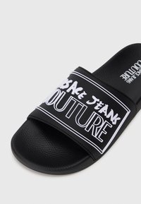 Fekete csúszós szandál, texturált gumitalppal és széles pánttal, amelyen fehér betűkkel olvasható a "VERSACE JEANS COUTURE" felirat, vastag betűtípussal.