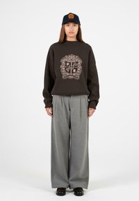 Hnědá oversized mikina se zdobným emblémem, kombinovaná s širokými šedými kalhotami. Model má na hlavě námořnickou čepici s oranžovým logem.