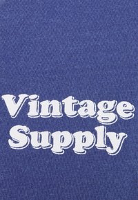 Niebieska koszulka z materiału, na której widnieje wyraźny, biały napis "Vintage Supply". Materiał wydaje się być miękki z teksturowanym wykończeniem.