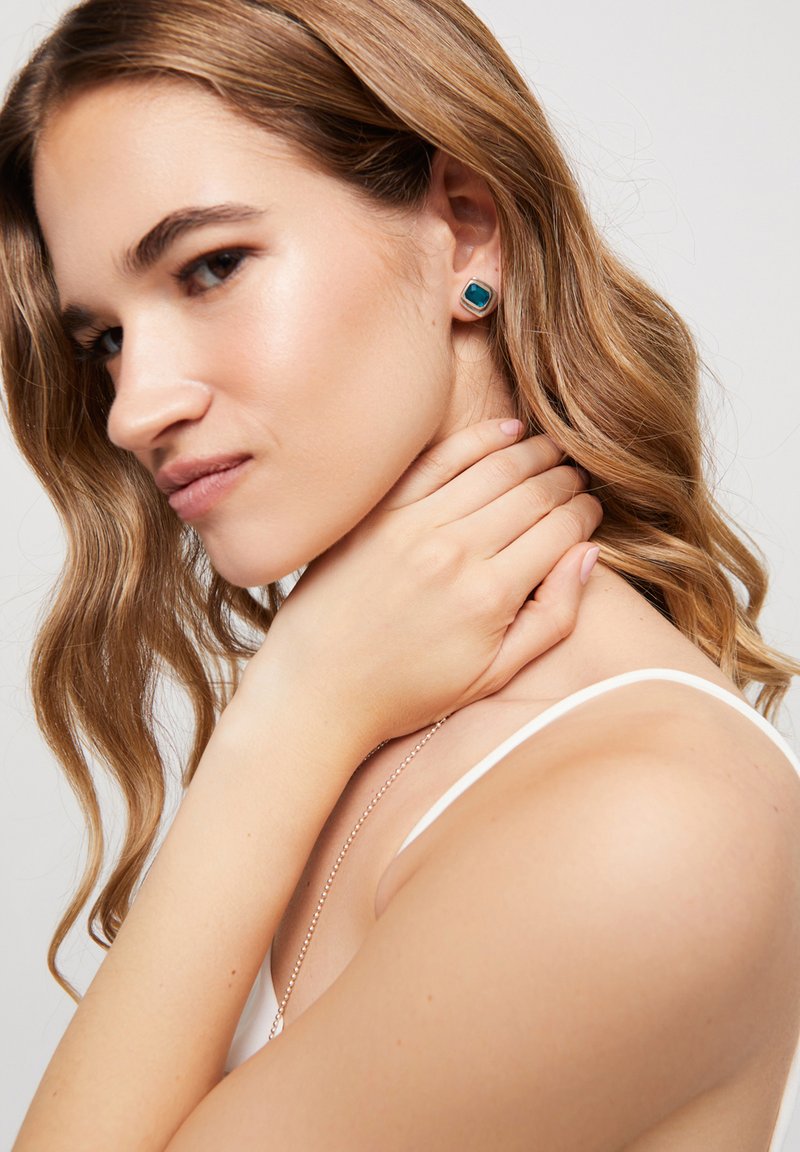 Pendientes cuadrados color verde azulado con borde metálico dorado. La modelo tiene el cabello largo y ondulado y se sujeta el cuello con una mano.
