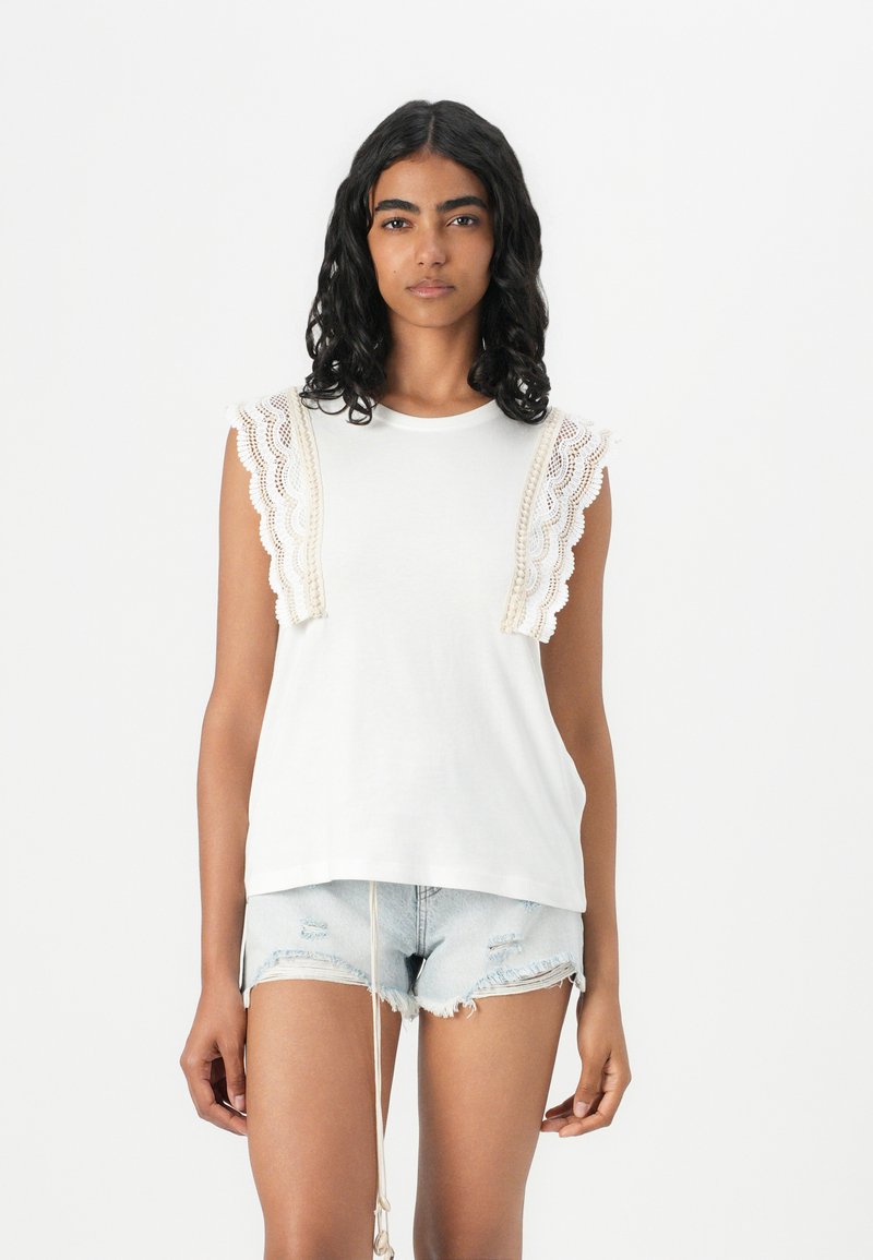 Top de algodón blanco sin mangas con detalles de encaje en los hombros, combinado con pantalones cortos de mezclilla desgastados en un tono azul claro, que presentan un cordón.