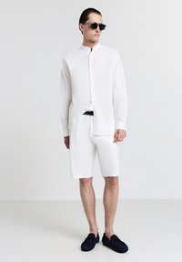 Camicia bianca a maniche lunghe con colletto alla mandarina, abbinata a pantaloni corti bianchi. Il modello indossa occhiali da sole neri e scarpe scivolate blu navy. Design minimalista.