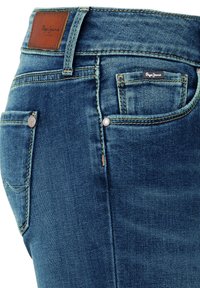 Sinine teksade lähivaade, kus on näha ees- ja tagataskud metallist neetidega, vööaas ning pruun nahast Pepe Jeansi logo.