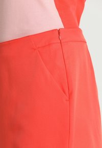 Koraliowe spodnie o gładkiej fakturze, z boczną kieszenią i dyskretnym zamkiem błyskawicznym. Materiał wygląda na lekki i miękko się układa.