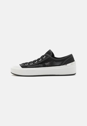 Svarte nett sneakers med en tykk hvit plattformsåle og sølvfarge aksent ved hælen, med svarte snøringer og hull.