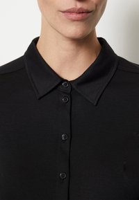 Svart pikétröja med klassisk krage, fem knappar, slät tygtextur och subtila sömnadsdetaljer vid axlarna.