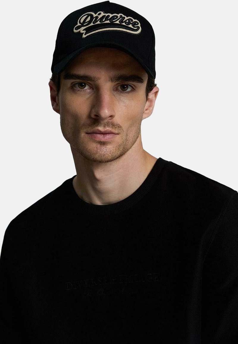 Czarna czapka z haftowanym logo "Diverse". Model nosi czarną bluzę z fakturowanej tkaniny i subtelnym brandingiem. Neutralne tło.