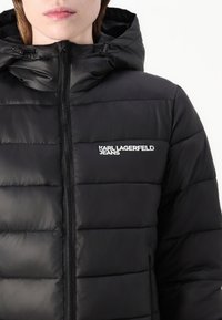 Juoda pūkinė striukė su kapišonu, horizontalaus siuvimo tekstūra, priekiniame užtrauktuke ir „Karl Lagerfeld Jeans“ logotipu kairėje krūtinės pusėje.