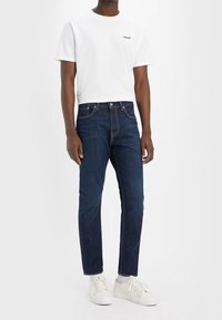 Vit kortärmad t-shirt med en liten svart Levi's-logotyp, tillsammans med mörkblå jeans och vita sneakers. Slät tyg och avslappnad passform.