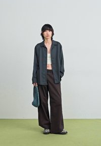 Överdimensionerad svart knapp-up skjorta, kombinerad med en vit crop-top, bruna vida byxor och randiga skor; modellen håller en blå väska.